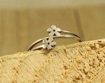 May include: A silver-colored ring featuring two small, stylized flower designs. The ring has a delicate, open design with the flowers positioned on either side of the band. The ring is displayed on a textured wooden surface.