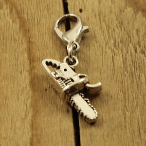 May include: A silver chainsaw charm with a lobster clasp. The charm features a detailed saw blade and engine parts. The background is a wooden surface with visible grain.