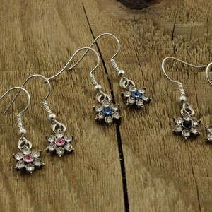May include: Three pairs of silver dangle earrings with flower-shaped charms. The flowers have clear rhinestones with a single pink, blue, or black rhinestone in the center.