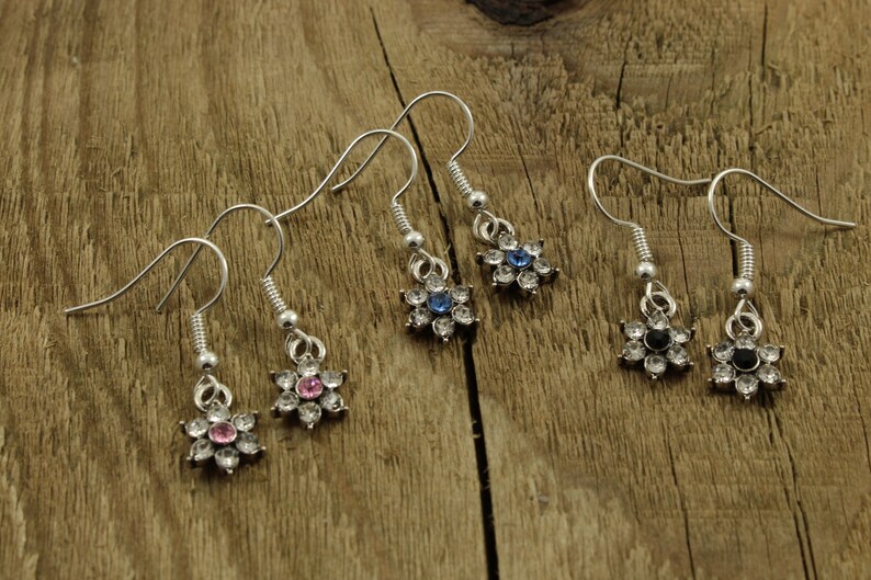 May include: Three pairs of silver dangle earrings with flower-shaped charms. The flowers have clear rhinestones with a single pink, blue, or black rhinestone in the center.