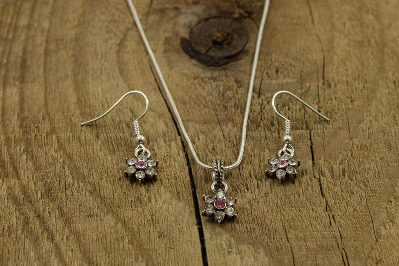 May include: A silver necklace and earrings set with a flower pendant and matching earrings. The flower pendant and earrings have clear and pink rhinestones.
