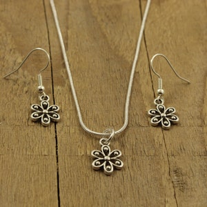 May include: A silver-tone jewellery set comprising a necklace with a flower pendant and two matching flower earrings. The floral design has six petals and a central bead. The set is presented on a wooden surface.