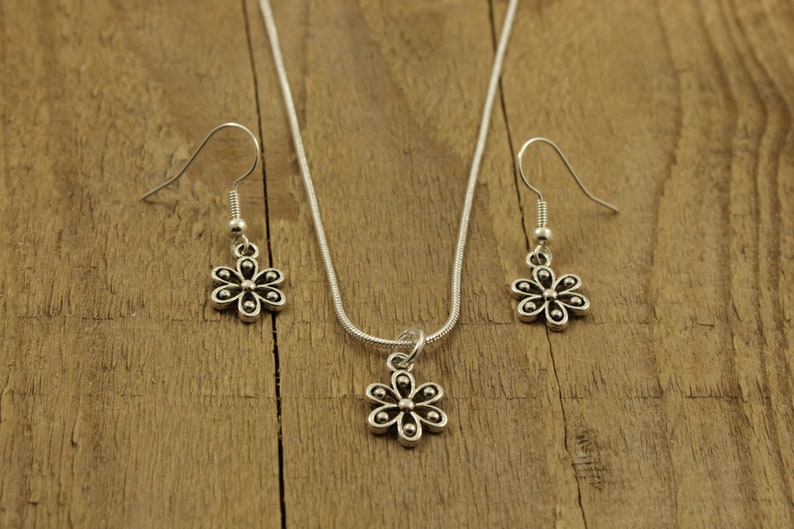 May include: A silver-tone jewellery set comprising a necklace with a flower pendant and two matching flower earrings. The floral design has six petals and a central bead. The set is presented on a wooden surface.