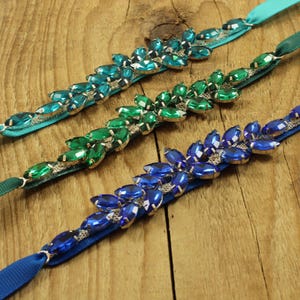 May include: Three satin ribbon garters with rows of jewel-toned rhinestones. The garters are in shades of teal, green, and blue.