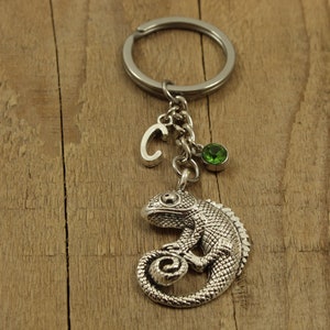 May include: Silver charm keychain featuring a detailed chameleon design with a green gemstone and a silver letter 'C' charm.