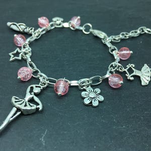 Puede incluir: Un brazalete de plata con cuentas rosas y varios dijes, incluyendo una bailarina, una estrella, una flor y un zapato.