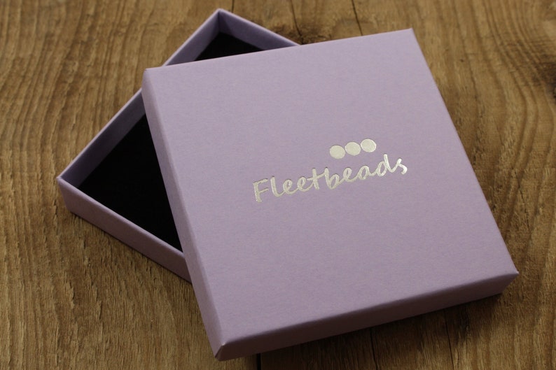 May include: A lavender colored gift box with a black interior. The box has a silver foil logo that says "Fleetbeads" with three silver circles above the text.