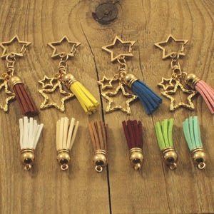 May include: A selection of star-shaped keyrings with gold-coloured metal fixings. Each keyring has a tassel in various colours, including brown, yellow, blue, and pink. The keyrings are displayed on a wooden surface.