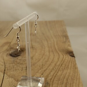 May include: A pair of silver star earrings with a simple design. The earrings are hanging from a clear display stand.