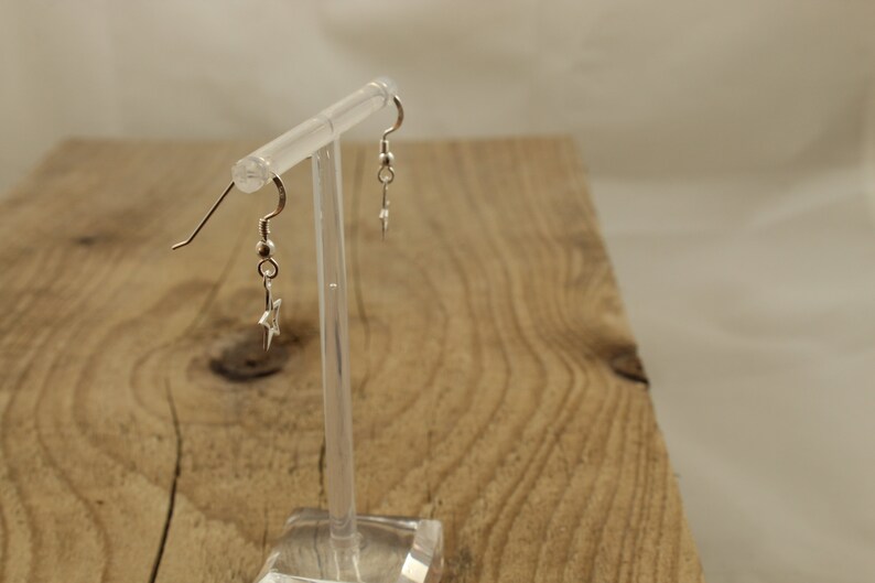 May include: A pair of silver star earrings with a simple design. The earrings are hanging from a clear display stand.