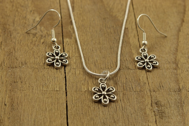 May include: A silver-coloured jewellery set featuring a daisy pendant necklace and matching earrings. The daisy pendants and earrings have six petals and small ball accents. The jewellery is displayed on a rustic wooden surface.