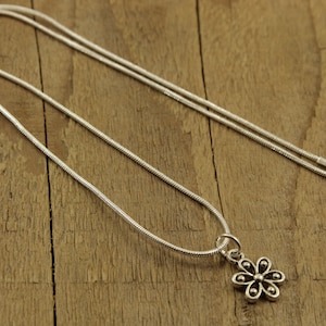 May include: A silver necklace featuring a flower pendant. The pendant has six petals and a small bead in the centre of each. The chain is a thin, snake chain style. The necklace is displayed on a wooden surface.