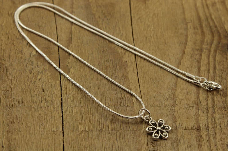 May include: A silver necklace featuring a flower pendant. The pendant has six petals and a small bead in the centre of each. The chain is a thin, snake chain style. The necklace is displayed on a wooden surface.