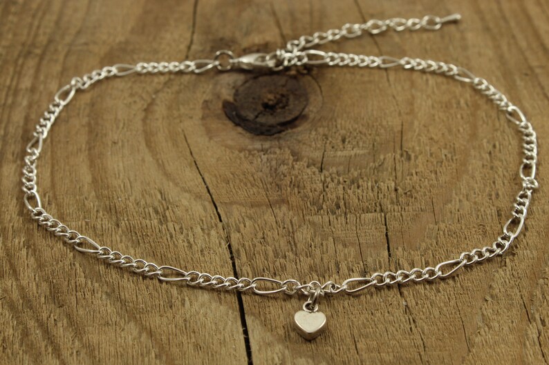 May include: A silver chain anklet with a small heart charm. The anklet is on a wooden surface.