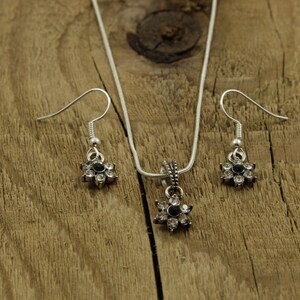 May include: A silver necklace and earrings set with a flower pendant and matching earrings. The flower pendant and earrings have a black center stone and clear rhinestones around the edges.