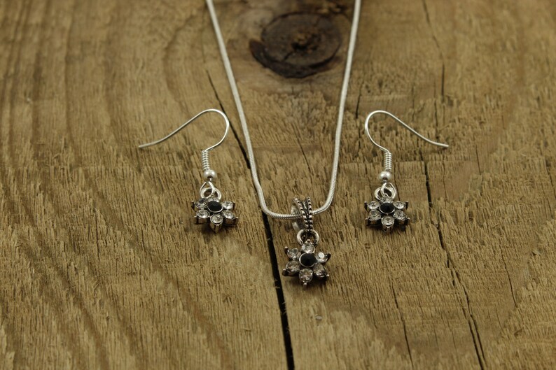 May include: A silver necklace and earrings set with a flower pendant and matching earrings. The flower pendant and earrings have a black center stone and clear rhinestones around the edges.
