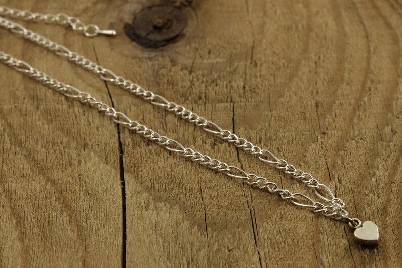 May include: A silver chain necklace with a small heart charm pendant. The chain is a figaro style with alternating links.