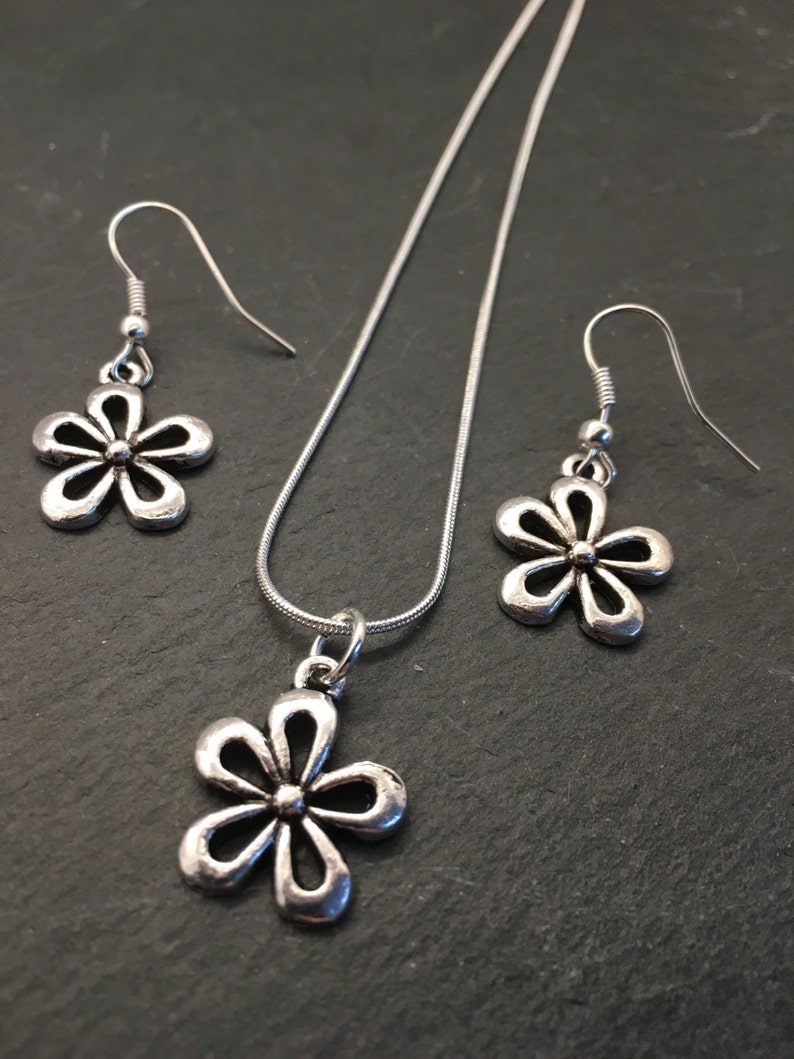 Pu&ograve; includere: Un set di collana e orecchini in argento con un delicato ciondolo a forma di fiore e orecchini abbinati. La collana ha una catena in argento e il ciondolo a forma di fiore ha un design semplice ed elegante.