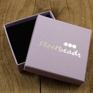 May include: A lavender-colored gift box with a black velvet interior. The box is open and the lid has the text "Fleetbeads" printed in silver foil.
