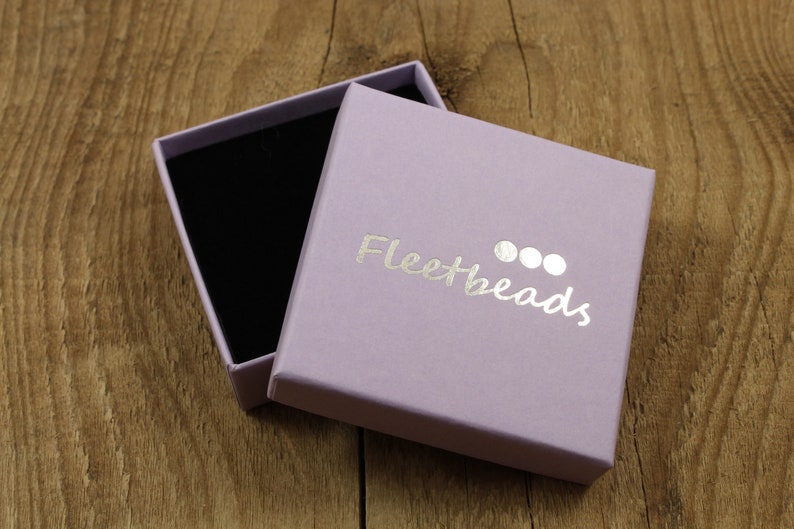 May include: A lavender-colored gift box with a black velvet interior. The box is open and the lid has the text "Fleetbeads" printed in silver foil.