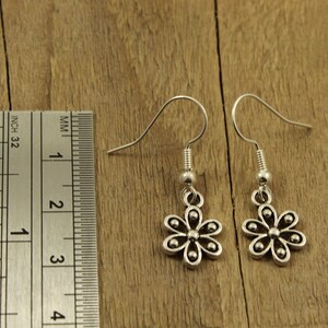 May include: A pair of silver flower earrings with hook fastenings. The earrings are shown on a wooden surface next to a ruler. Each earring has a six-petal flower design with small, round details. The ruler shows inches and millimeters.