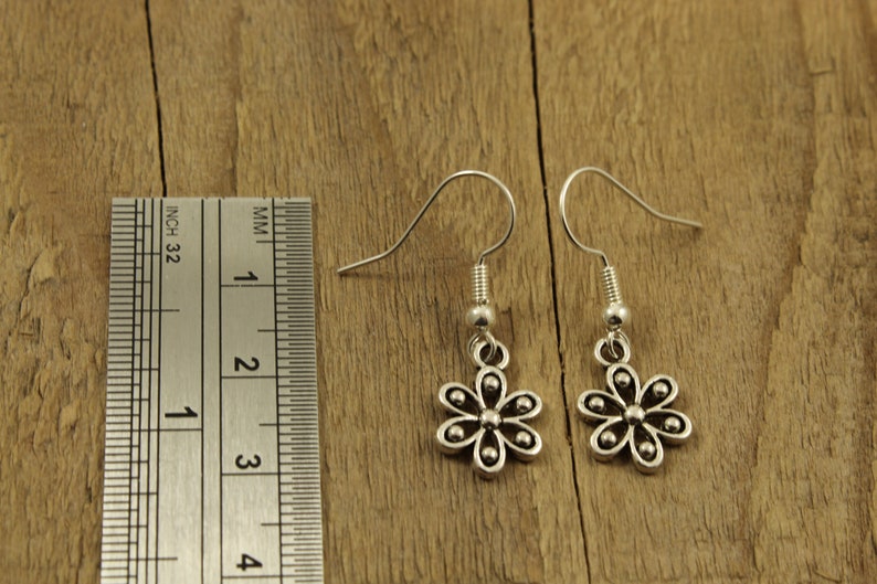 May include: A pair of silver flower earrings with hook fastenings. The earrings are shown on a wooden surface next to a ruler. Each earring has a six-petal flower design with small, round details. The ruler shows inches and millimeters.