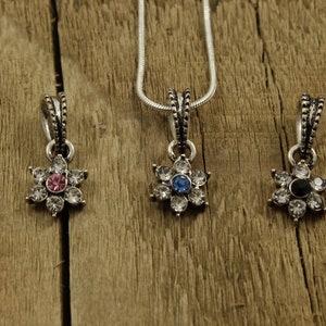 May include: Three silver flower-shaped pendants with clear crystal accents. The center stone of each pendant is a different color: pink, blue, and black.