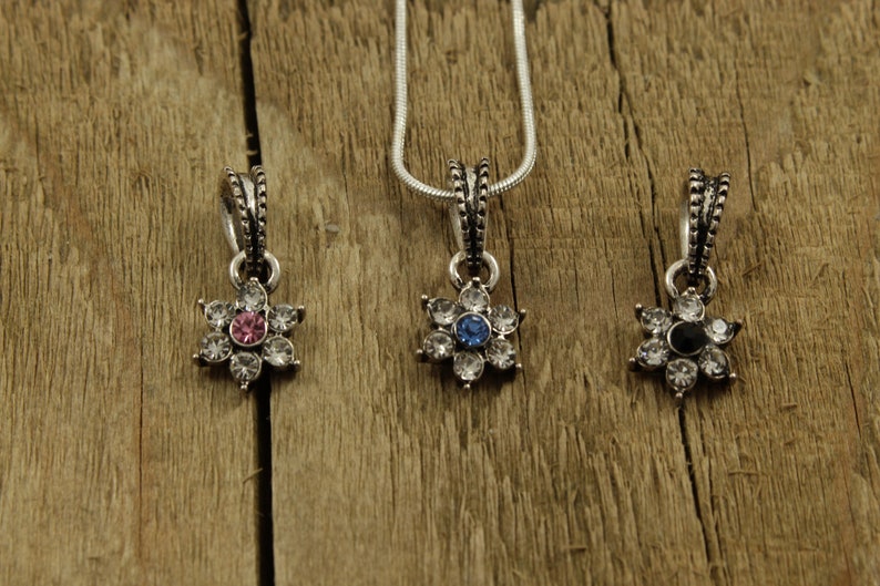 May include: Three silver flower-shaped pendants with clear crystal accents. The center stone of each pendant is a different color: pink, blue, and black.