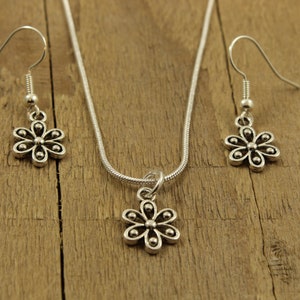 May include: A silver-toned jewellery set comprising a necklace and earrings. The necklace features a flower pendant on a fine chain. The earrings, also flower-shaped, dangle from hooks. The set is presented on a wooden surface.