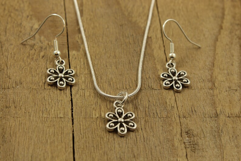 May include: A silver-toned jewellery set comprising a necklace and earrings. The necklace features a flower pendant on a fine chain. The earrings, also flower-shaped, dangle from hooks. The set is presented on a wooden surface.