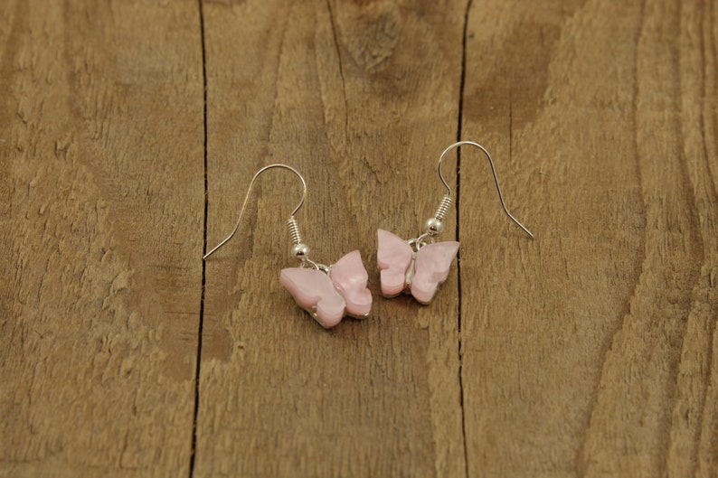 May include: A pair of silver butterfly earrings with pink butterfly charms. The earrings are hanging from a wooden surface.