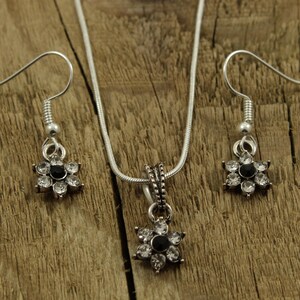 May include: A silver necklace and earrings set with a flower pendant and matching earrings. The flower pendant and earrings have a black center stone surrounded by clear rhinestones.