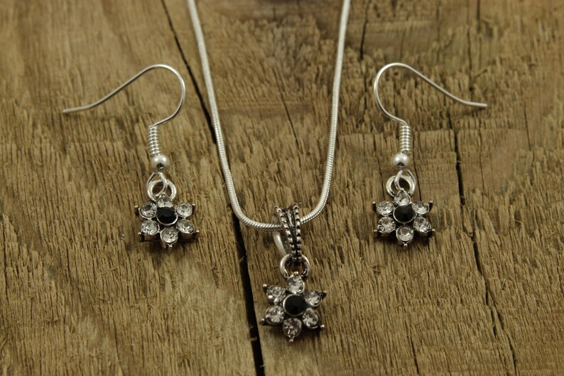 May include: A silver necklace and earrings set with a flower pendant and matching earrings. The flower pendant and earrings have a black center stone surrounded by clear rhinestones.