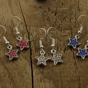May include: A set of six star-shaped dangle earrings with crystal accents. The earrings are silver-toned and feature red, blue, and clear crystals.