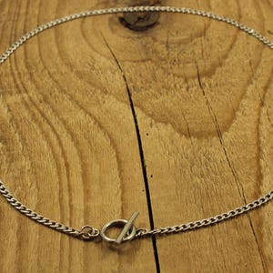 May include: A silver-toned metal chain necklace featuring a circular ring and a perpendicular bar toggle clasp. The necklace is formed by a series of small, interlocking links that create a continuous, uniform chain design around the entire piece.