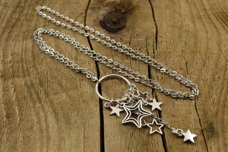Pode incluir: Um colar de corrente de prata com um pingente circular e v&aacute;rios pingentes de estrelas. O colar est&aacute; sobre uma superf&iacute;cie de madeira.