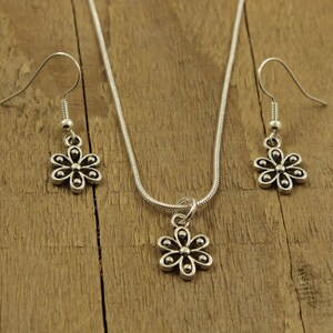 May include: A silver-coloured jewellery set featuring a daisy pendant necklace and matching drop earrings. The necklace has a fine chain, and the earrings have hook closures. The floral design is detailed with small, round accents.