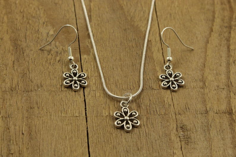 May include: A silver-coloured jewellery set featuring a daisy pendant necklace and matching drop earrings. The necklace has a fine chain, and the earrings have hook closures. The floral design is detailed with small, round accents.
