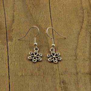 May include: A pair of silver flower earrings with hook fastenings. The earrings feature a small, detailed floral design. The background is a wooden surface.