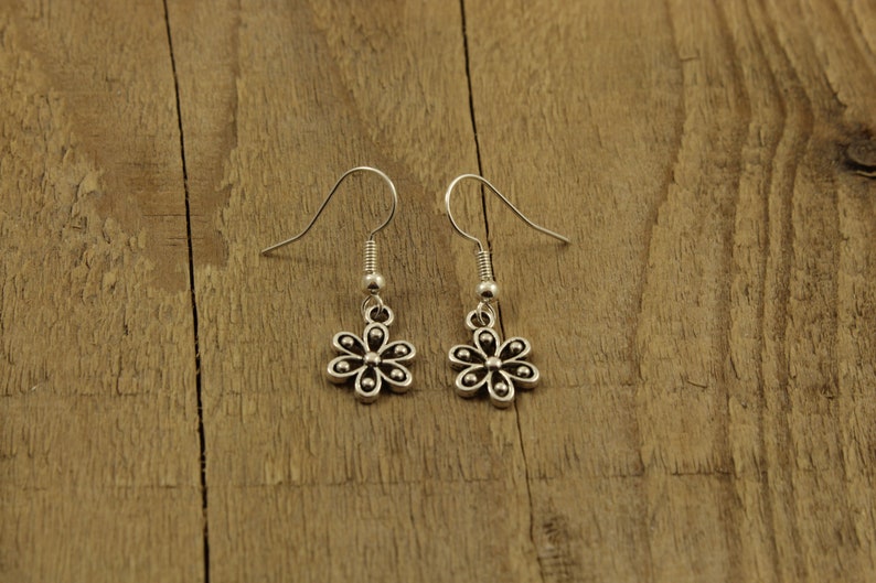 May include: A pair of silver flower earrings with hook fastenings. The earrings feature a small, detailed floral design. The background is a wooden surface.