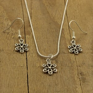 May include: A silver-coloured jewellery set featuring a flower design. The set includes a necklace with a pendant and a pair of matching earrings. The jewellery is displayed on a wooden surface.
