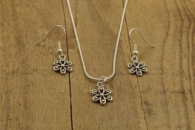 May include: A silver-coloured jewellery set featuring a flower design. The set includes a necklace with a pendant and a pair of matching earrings. The jewellery is displayed on a wooden surface.