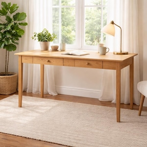 May include: A light wood desk with three drawers, a brass lamp, and a white chair. The desk is in front of a window with sheer curtains. A woven rug and a large potted plant are in the room.