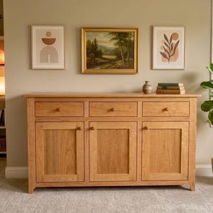 May include: A light brown wooden sideboard featuring three drawers positioned above three cabinet doors, each equipped with a small round knob. The piece is constructed from wood with a visible grain and sits on a carpeted floor.