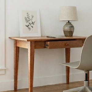 May include: A wooden desk with four tapered legs and a single pull-out drawer featuring a small round knob. On the desk sits a table lamp with a beige fabric shade and a ceramic base, a small book, and a framed botanical line art print.