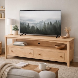 May include: A light wood TV stand with two drawers and a shelf, holding a flat-screen TV displaying a misty forest scene. A small plant, books, and decorative bowls are on the stand. A lamp and a floor lamp are also visible.