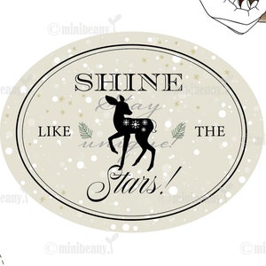 Könnte beinhalten: Ein beigefarbenes ovales Schild mit einer schwarzen Silhouette eines Rehs mit Schneeflocken darauf. Das Schild sagt "Shine Like The Stars!" mit einem goldenen Sternenmuster im Hintergrund.