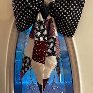 May include: A patriotic door bow featuring a large bow with a black background and white stars. The bow's tails display a patchwork design in red, white, and blue, creating a festive and decorative accent for the front door.
