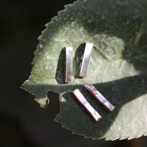 May include: Four silver bar earrings are arranged on a green leaf. The earrings have a rectangular shape with a hammered texture. The leaf is a deep green color with some imperfections and is set against a dark background.