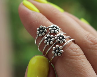 Könnte beinhalten: Ein silberner Ring mit mehreren Bändern, die mit kleinen Blumenmotiven verziert sind. Der Ring wird an einem Finger mit gelbem Nagellack getragen.